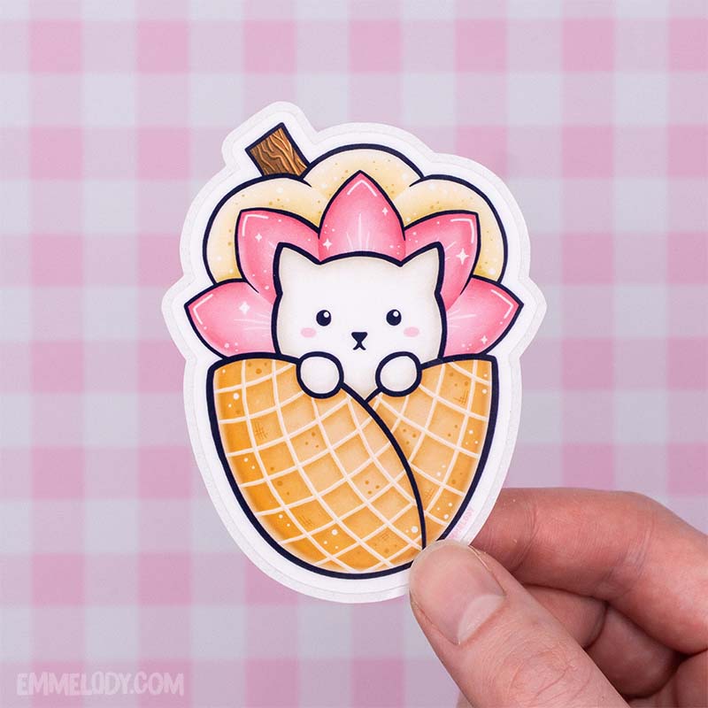 Glossy vinyl sticker by EmMelody showing a kawaii illustration of a white cat tucked inside an ice cream cone, held in front of a pink gingham background