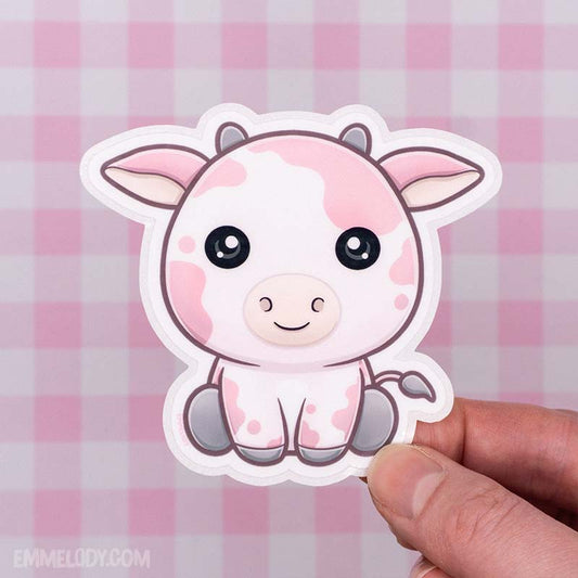 Glossy vinyl sticker by EmMelody showing a kawaii illustration of a sitting pink cow with splotches, held by a human hand in front of a pink gingham background.